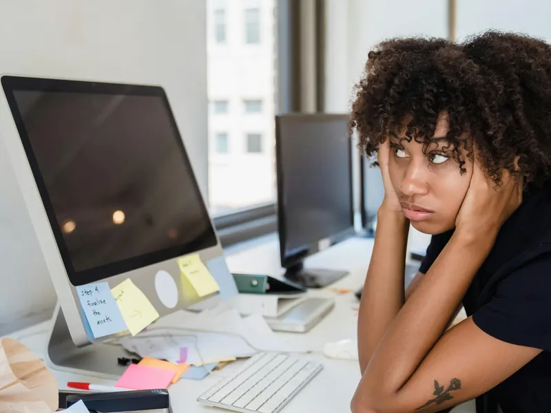 Mulher distraída diante do computador em uma mesa cheia de papéis, representando procrastinação